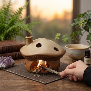 May include: A ceramic mushroom-shaped essential oil diffuser with a chimney and three holes on top. The diffuser is lit with a small flame inside. A hand is holding a bundle of herbs. A small bottle of essential oil and a cup are nearby.