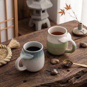 May include: Two ceramic mugs with whale tail handles. One mug is blue and white, the other is white and green. Both mugs are filled with a dark liquid. The mugs are on a wooden table with decorative items.