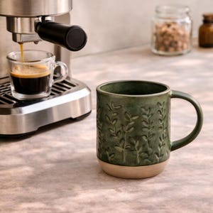 May include: A green ceramic mug with a beige base and a handle, decorated with a raised leaf pattern. Espresso is being poured into a clear glass cup from a stainless steel espresso machine. Jars are visible in the background.