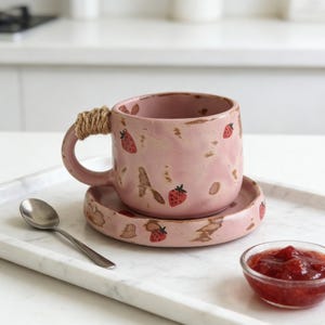 May include: Pink ceramic mug and saucer set decorated with red strawberries. The mug handle is wrapped with twine. A small glass bowl of strawberry jam and a silver spoon are also present.