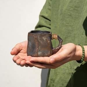 May include: A handcrafted, square-shaped ceramic mug with a dark, textured finish. The handle is wrapped with a natural fiber. The mug is held in open hands against a green shirt and white background. A green beaded bracelet is visible.