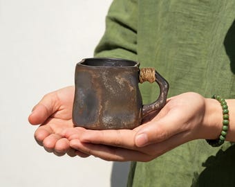 May include: A handcrafted, square-shaped ceramic mug with a dark, textured finish. The handle is wrapped with a natural fiber. The mug is held in open hands against a green shirt and white background. A green beaded bracelet is visible.