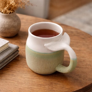May include: A ceramic mug with a whale tail handle. The mug is white at the top, transitioning to a light green base with a thin blue stripe. The mug is filled with a dark liquid, likely tea or coffee, and sits on a wooden table.