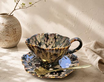 May include: A decorative teacup and saucer set with a unique speckled glaze in shades of brown, black, and gold. The cup has a wavy rim and a matching saucer. A gold spoon rests on the saucer, and a small vase with a sprig of greenery is in the background.