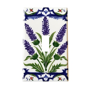 Puede incluir: Una placa de interruptor de luz de cerámica blanca con un diseño floral. El diseño incluye flores de lavanda moradas, hojas verdes y detalles decorativos azules. La abertura del interruptor es rectangular.