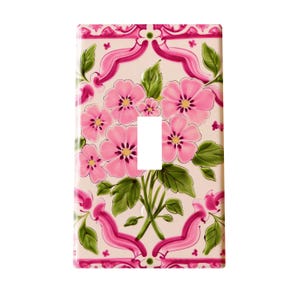 May include: A decorative light switch cover featuring a floral design. The cover has a rectangular shape with a central opening for the switch. Pink flowers with yellow centers and green leaves are arranged on a cream background, framed by pink ribbons.