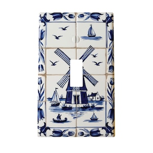 May include: A blue and white ceramic light switch cover with a Dutch windmill, houses, and boats. The switch cover is made of tiles and has a traditional Delft blue design.