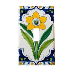 May include: A decorative ceramic light switch cover featuring a yellow daffodil with green leaves against a white background. The design is framed by a blue border with floral accents. The switch opening is rectangular.