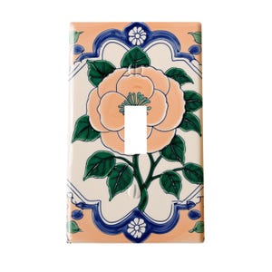 May include: A ceramic light switch cover featuring a large peach-colored flower with green leaves and a dark green stem. The flower is centered on a cream-colored background, framed by a blue and peach border with floral accents. The cover has a single switch opening.