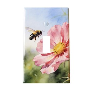 May include: A decorative light switch cover featuring a watercolor painting of a bee in flight near a pink flower with a yellow center. The background has soft green and blue hues.