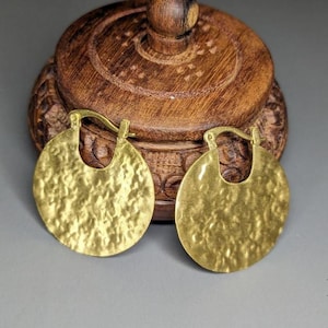 Può includere: Un paio di orecchini a cerchio color oro con texture martellata. Gli orecchini sono circolari con fondo piatto e chiusura a gancio. Sono esposti davanti a una scatola di legno intagliato con coperchio rotondo.