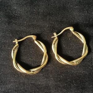 Op de afbeelding: Een paar goudkleurige oorringen met een gedraaid touwontwerp. De oorbellen liggen op een donkere, getextureerde ondergrond. Klassieke sieraden voor elke gelegenheid.