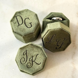 May include: Three olive green velvet ring boxes, each with a unique monogram in black script. One box is open, revealing a silver ring. The boxes are octagonal and have a soft, textured appearance.