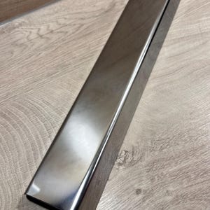 Könnte beinhalten: Ein rechteckiger Metallgegenstand mit gebürsteter Oberfläche und polierter Oberfläche. Das Metall hat eine silberne Farbe und reflektiert das Licht. Das Objekt ist wahrscheinlich ein Bestandteil einer größeren Struktur oder eines Möbelstücks. Die Kanten sind leicht abgerundet.