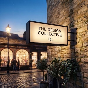 May include: A rectangular, illuminated, double-sided projecting sign mounted on a brick wall. The sign features a white face with black text reading THE DESIGN COLLECTIVE and a DC logo. The sign is positioned above a cobblestone street.