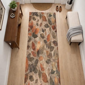 Puede incluir: Una alfombra rectangular con un diseño de hojas en tonos marrón, naranja y gris. La alfombra está colocada en una entrada, con una consola de madera, un banco y una puerta en el fondo. La alfombra mide aproximadamente 91 cm de ancho y 183 cm de largo.
