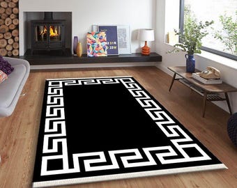 May include: A rectangular black rug with a white Greek key border. The rug is placed on a wooden floor in a room with a fireplace and a window. The rug has a fringe on the short ends.