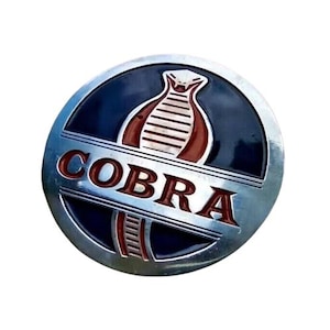 May include: A round, silver-colored emblem with the word "COBRA" in red lettering. The emblem features a stylized cobra design in red and white against a dark blue background. The emblem has a metallic finish.