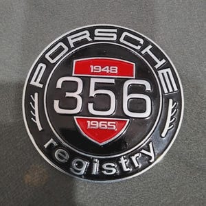 May include: A round, black and silver Porsche 356 registry emblem. The emblem features the word "Porsche" in white lettering around the outer edge. The number "356" is prominently displayed in the center, with the years 1948 and 1965.