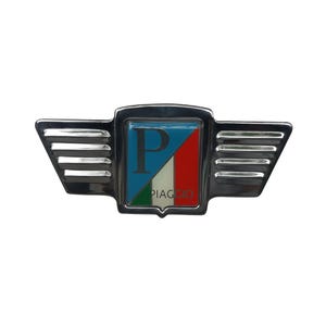Puede incluir: Un emblema Piaggio cromado con un centro en forma de escudo que muestra la letra "P" en azul, blanco y rojo, y la palabra "PIAGGIO". El emblema tiene extensiones en forma de alas a ambos lados.
