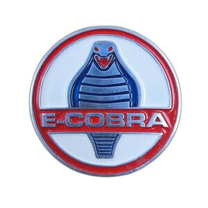 May include: A round, metallic emblem with a red border and the text "E-COBRA" in white. The emblem features a blue cobra design with a red open mouth, set against a white background.