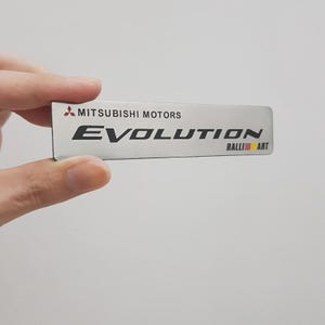 Könnte beinhalten: Ein rechteckiges Emblem aus silberfarbenem Metall mit dem Mitsubishi Motors Logo, dem Wort EVOLUTION in großen schwarzen Buchstaben und dem RALLIART Logo mit roten und gelben vertikalen Streifen auf der rechten Seite vor einem schlichten Hintergrund.