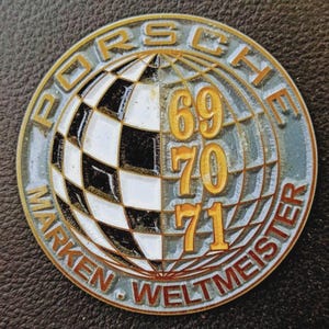 Puede incluir: Un emblema Porsche redondo y vintage con borde dorado y las palabras "PORSCHE MARKEN WELTMEISTER". Presenta un diseño de globo a cuadros con los números "69, 70, 71" en dorado.
