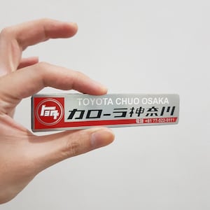 Może przedstawiać: Prostokątny srebrny identyfikator z czerwonym kwadratem po lewej stronie, zawierającym białe logo Toyota. Tekst brzmi TOYOTA CHUO OSAKA, po którym następują japońskie znaki oraz numer telefonu +81 77-522-9111 w kolorze czarno-białym.