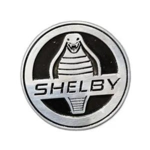 May include: A circular, silver-colored Shelby emblem with a black background. The emblem features a cobra design above the word "SHELBY" in capital letters. The cobra is detailed with scales and a menacing expression.