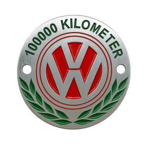 May include: A circular, silver metal emblem with the text "100000 KILOMETER" in green. It features a red "VW" logo at the centre, encircled by a green laurel wreath. Two small holes are visible on the sides of the emblem.