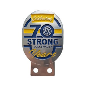 Peut inclure: Un emblème métallique argenté et bleu célébrant les 70 ans de Volkswagen. L'emblème comporte les textes "Celebrating", "70", "STRONG", "Volkswagen" et "Years" en jaune et blanc. Il a une base rectangulaire avec deux trous.