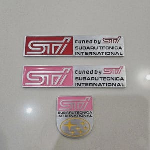 May include: Three rectangular and shield-shaped metal emblems featuring the STI logo. Two rectangular emblems display the text tuned by STI SUBARU TECNICA INTERNATIONAL in black, with one featuring a red background and one a pink background. The shield emblem is pink with the STI logo and SUBARU TECNICA INTERNATIONAL text above a yellow star cluster.