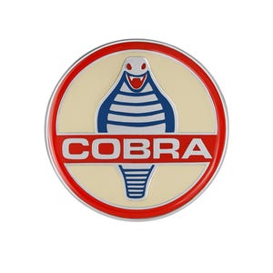 May include: A round emblem with a red border and the word "COBRA" in white letters. Inside, a cream-colored background features a blue and white cobra design.