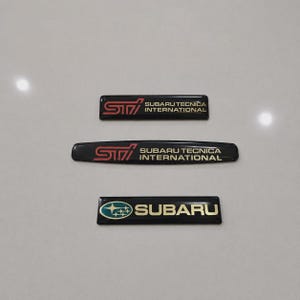 May include: Three rectangular black emblems with glossy finishes. Two emblems feature the red STI logo and the text SUBARU TECNICA INTERNATIONAL in gold. The third emblem displays the blue and silver Subaru star logo with the word SUBARU in gold.
