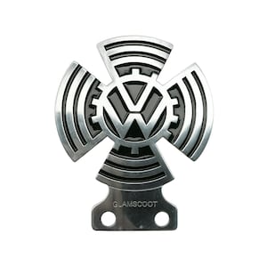Puede incluir: Un emblema metálico plateado con el logotipo de Volkswagen en el centro, rodeado por un diseño en forma de cruz con detalles de engranajes. El emblema tiene una base rectangular con dos agujeros y el texto "GLAMSCOOT".