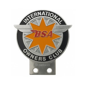 May include: A silver-colored metal emblem with the text "INTERNATIONAL OWNERS CLUB" in a circular design. The emblem features a central orange starburst with the red letters "BSA". The bottom of the emblem has the text "GLAMSCOOT".