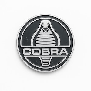 Könnte beinhalten: Ein rundes, schwarz-silbernes Emblem mit einem Kobra-Design. Das Wort "COBRA" ist in silbernen Buchstaben unter dem Kobra-Bild dargestellt. Das Emblem hat einen silbernen Rand und ist vor einem weißen Hintergrund platziert.