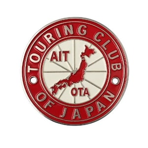 Può includere: Una spilla rotonda, rossa e bianca con la scritta "TOURING CLUB OF JAPAN" sul bordo. Il centro presenta una mappa del Giappone in rosso, con le scritte "AIT" e "OTA".