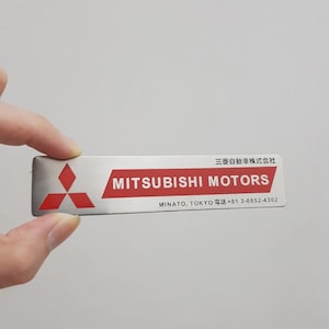 May include: A rectangular silver metal nameplate featuring the red Mitsubishi Motors logo on the left and the text MITSUBISHI MOTORS in white on a red background. It includes Japanese text, MINATO, TOKYO, and the phone number +81 3-6852-4302.