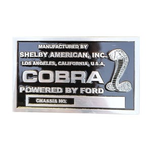 以下が含まれることがあります： 黒い背景とシルバーのトリムが付いた長方形の金属製エンブレム。エンブレムには「COBRA POWERED BY FORD」と「SHELBY AMERICAN, INC.」の文字とコブラのエンブレムが描かれています。「CHASSIS NO.」の文字も表示されています。