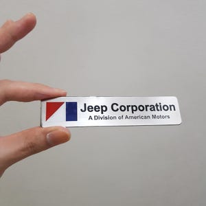 Könnte beinhalten: Ein rechteckiges Emblem aus silbernem Metall mit einem rot-blauen geometrischen Logo auf der linken Seite. Der Text lautet Jeep Corporation, gefolgt von dem kleineren Text A Division of American Motors in schwarzer Schrift auf silbernem Grund.