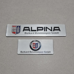 Puede incluir: Dos emblemas rectangulares de metal plateado con el logotipo y el texto de Alpina. El emblema superior muestra el escudo de Alpina y el texto ALPINA Burkard Bovensiepen GmbH. El emblema inferior presenta un logotipo circular de Alpina y el texto Burkard Bovensiepen GmbH.