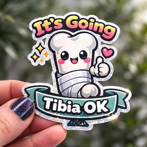 May include: A sticker with the text "It's Going" above a cartoon chef's hat character with a bandaged leg, and the text "Tibia OK" on a banner. The character is giving a thumbs up. The sticker has a glittery border.