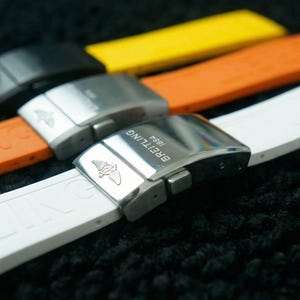 Könnte beinhalten: Drei Kautschuk-Uhrenarmbänder in Weiß, Orange und Gelb, jeweils mit einer metallischen Faltschließe. Die weißen und orangefarbenen Armbänder sind mit dem Schriftzug BREITLING 1884 und einem geflügelten Logo auf der silberfarbenen Schließe versehen.