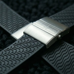 Puede incluir: Primer plano de una correa de reloj negra con un cierre de acero inoxidable. La correa tiene un diseño de patrón de ondas. El cierre está grabado con el nombre de la marca "ULYSSE NARDIN".