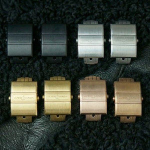 May include: An assortment of eight watch clasps in various metallic finishes, including black, silver, and gold tones. The clasps are arranged in two rows, showcasing different color options and designs. The clasps are on a textured black surface.