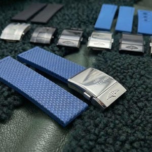 Puede incluir: Conjunto de correas y cierres de reloj en varios colores. Incluye componentes azules, grises y plateados. Las correas azules tienen un patrón texturizado. Los cierres son plateados con el logo "Breitling 1884".