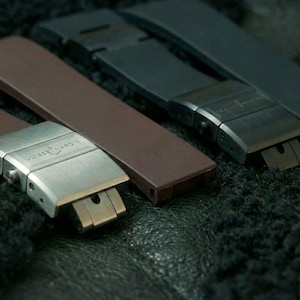 May include: Close-up of three watch straps in shades of brown and grey. The straps have metal clasps with the brand name engraved. The watch straps are lying on a dark, textured surface.