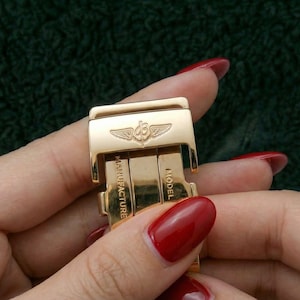 May include: Close-up of a gold-colored watch clasp with the brand logo and the words "Manufacture" and "Model" engraved. The clasp is held in a hand with red nail polish against a dark green background.