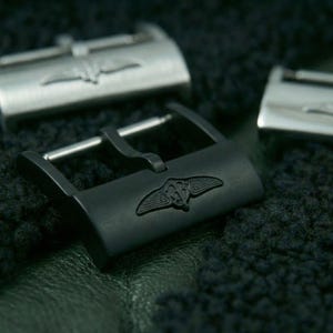 May include: Close-up of three metal watch buckles. One is black with a winged emblem, and two are silver with the same emblem. The buckles are on a dark, textured surface.